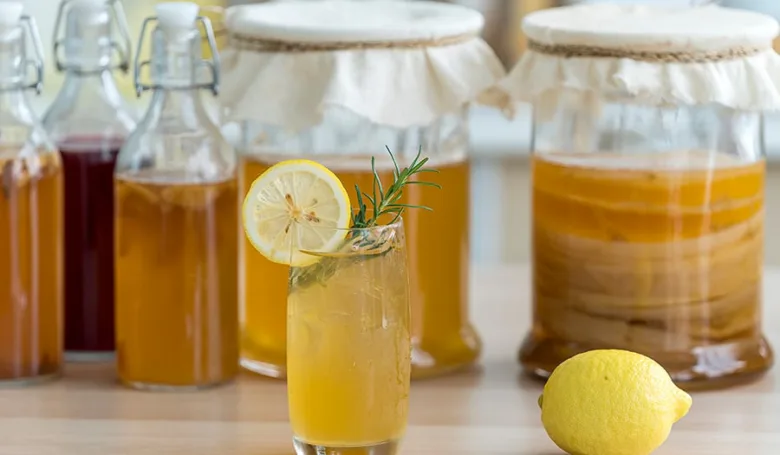 Sklenice kombuchy s citronem a růžmarýnem před nádobami na fermentaci
