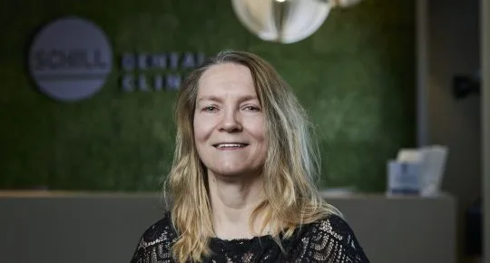 Žena usmívá v ordinaci zubního lékaře s logem kliniky na pozadí.