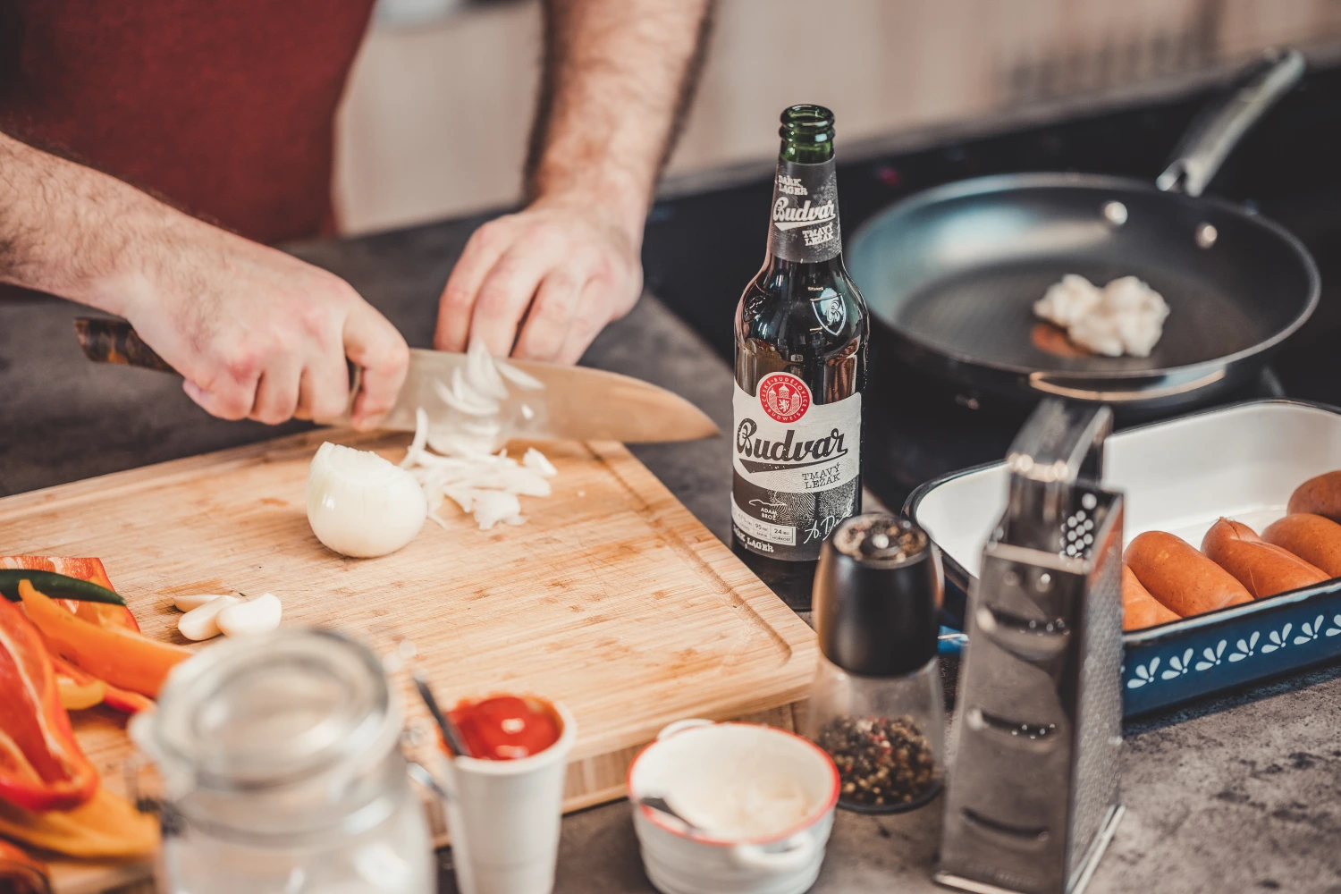 Příprava buřtů na pivu – nakrájená cibule, pivo Budvar a klobásy
