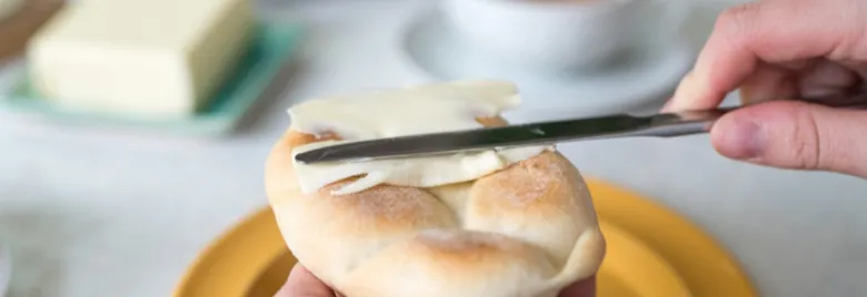 Ruka rozmazává čerstvé máslo na teplém rohlíku.