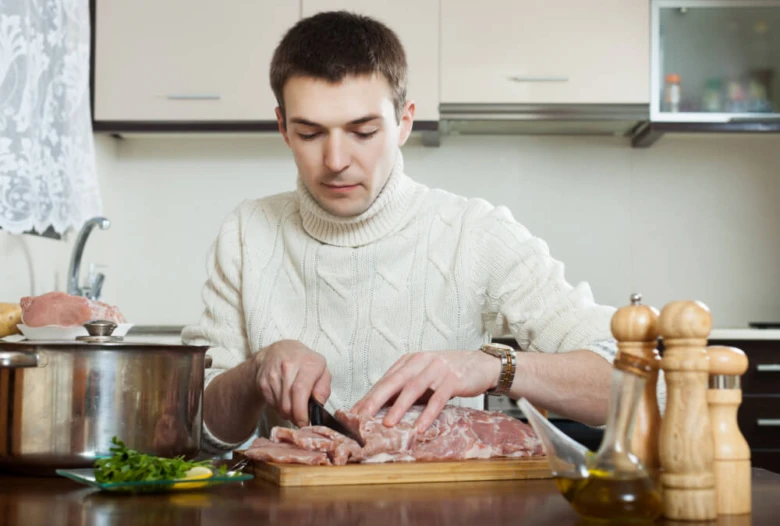 Muž řezá surové maso na dřevěném prknu v kuchyni.