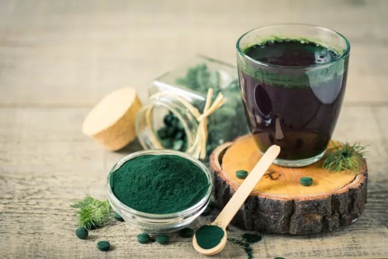 Sklenička s tmavě zelenou spirulinou nebo chlorellou vedle prášku a kapslí.