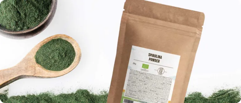 Zelený prášek spiruliny v lžičce vedle balení.
