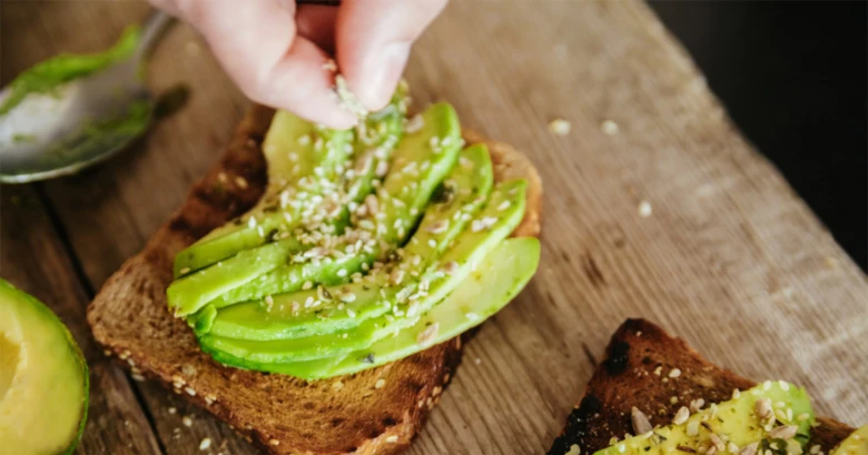 Ruce posypávají semínka na toast s nakrájeným avokádem.